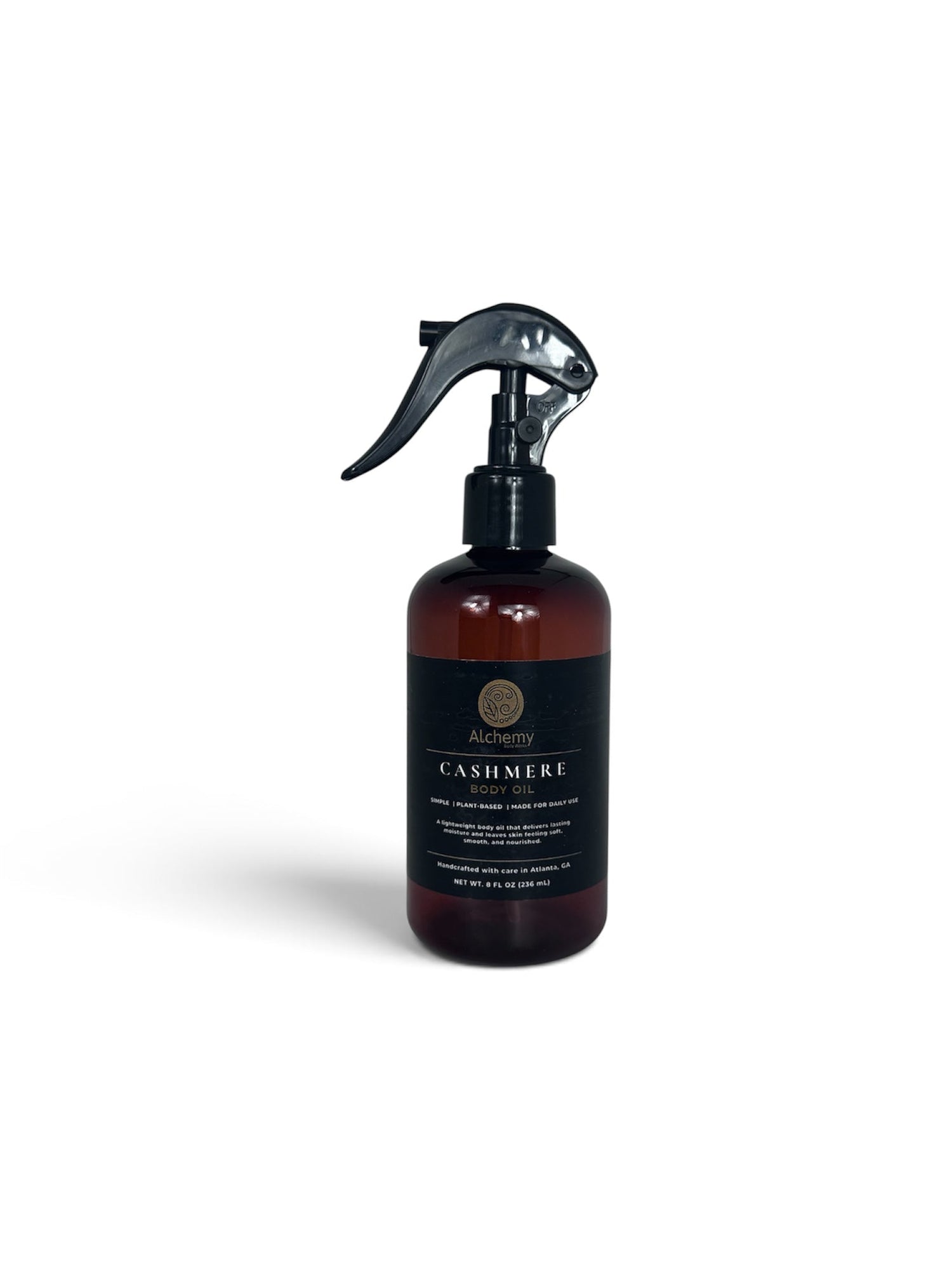 An amber spray bottle with a black trigger sprayer and a black label of Alchemy Body Werks Cashmere Body Oil on a clean white background.