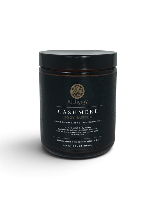 A amber jar with black lid and black label of Alchemy Cashmere Body Butters on a white background with a soft shadow.