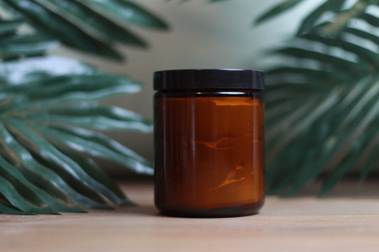 Brown glass jar with black lid on a wooden surface with green leaves in the background contains creme body butter