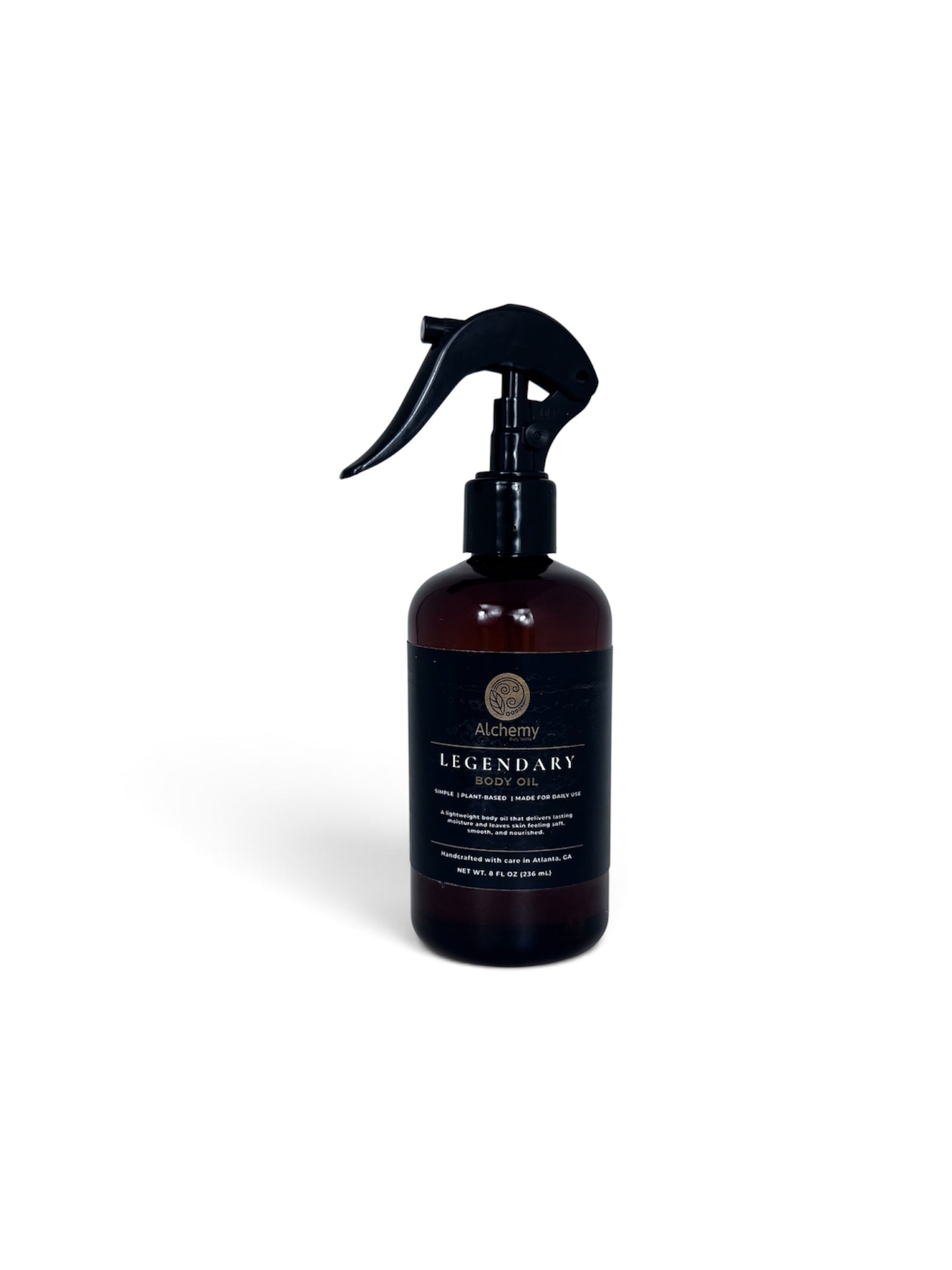 An amber spray bottle with a black trigger sprayer and a black label of Alchemy Body Werks Legendary Body Oil on a soft white background.
