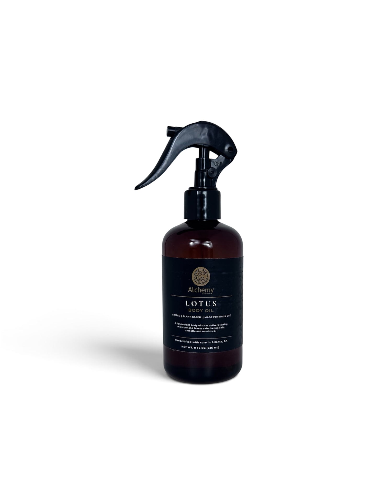 An amber spray bottle with a black trigger sprayer and a black label of Alchemy Body Werks Lotus Body Oil on a soft white background.