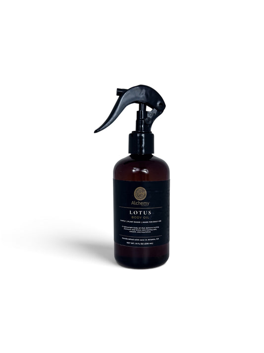 An amber spray bottle with a black trigger sprayer and a black label of Alchemy Body Werks Lotus Body Oil on a soft white background.