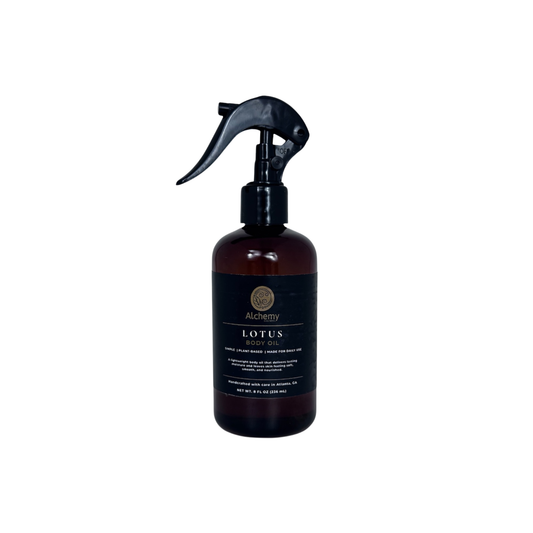 An amber spray bottle with a black trigger sprayer and a black label of Alchemy Body Werks Lotus Body Oil on a white background.