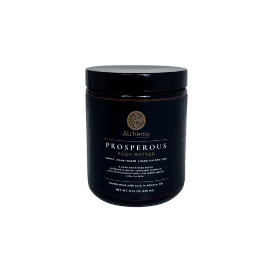 An amber jar with a black lid and black label of Alchemy Body Werks Prosperous Body Butter on a white background.