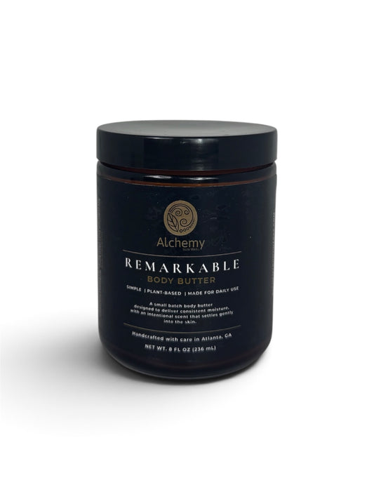 An amber jar with a black lid and black label of Alchemy Body Werks Remarkable Body Butter on a white background with a soft shadow.